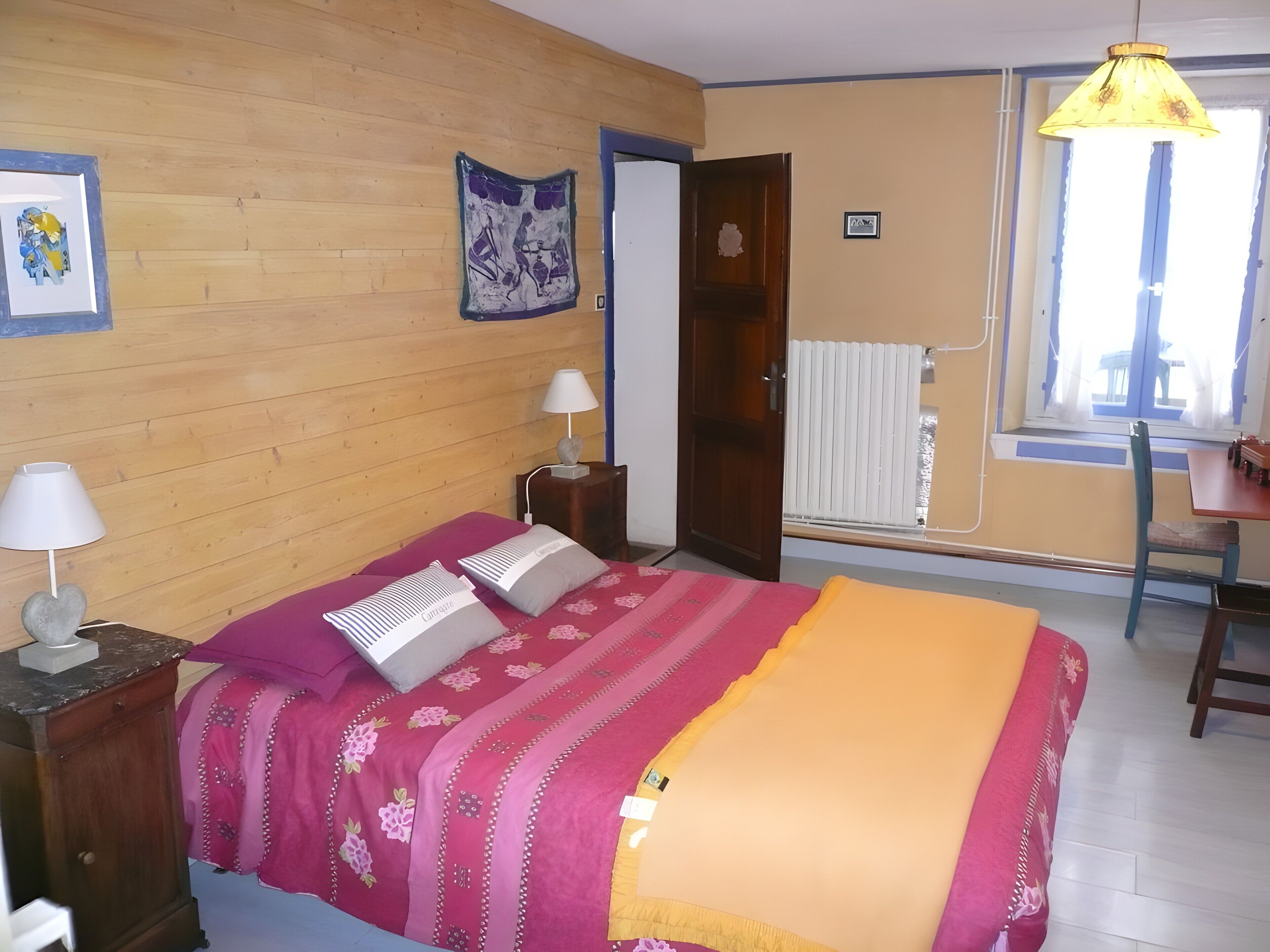 A bedroom with a wooden wall, a bed with a pink and yellow blanket, a nightstand with a lamp, and a window with a view.