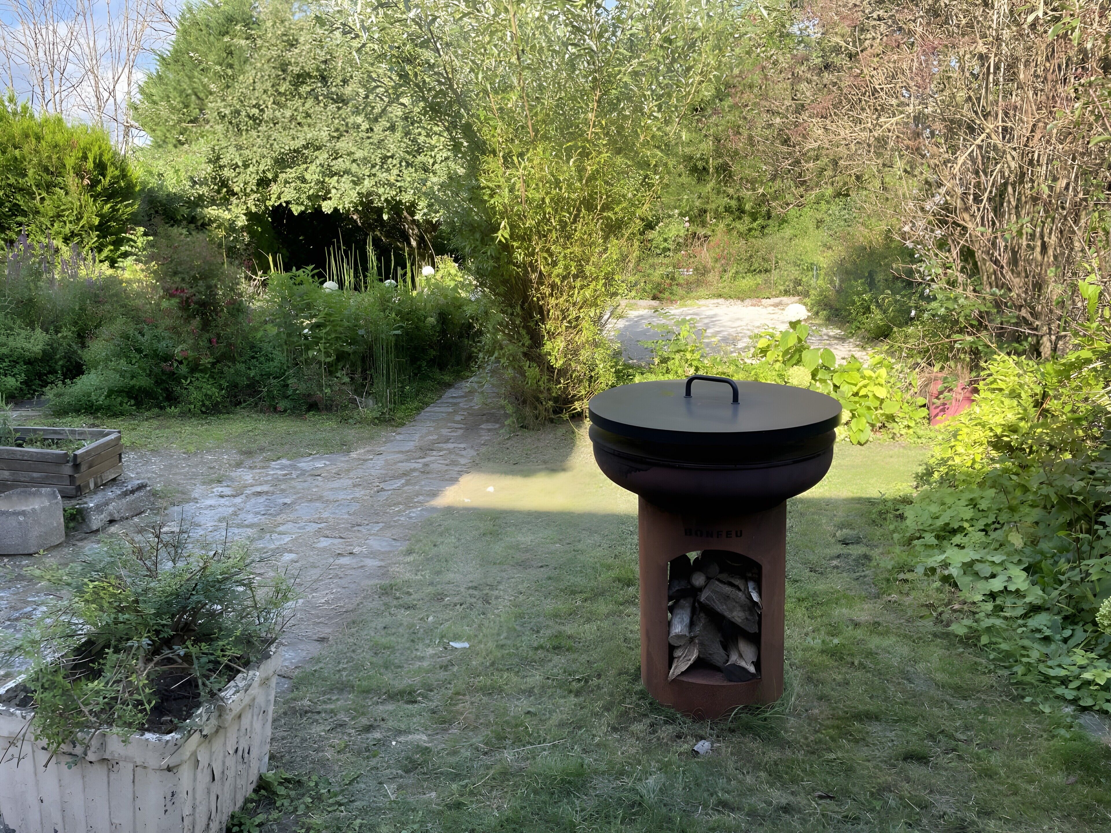 A barbecue grill in a garden with a stone path, surrounded by greenery and trees.