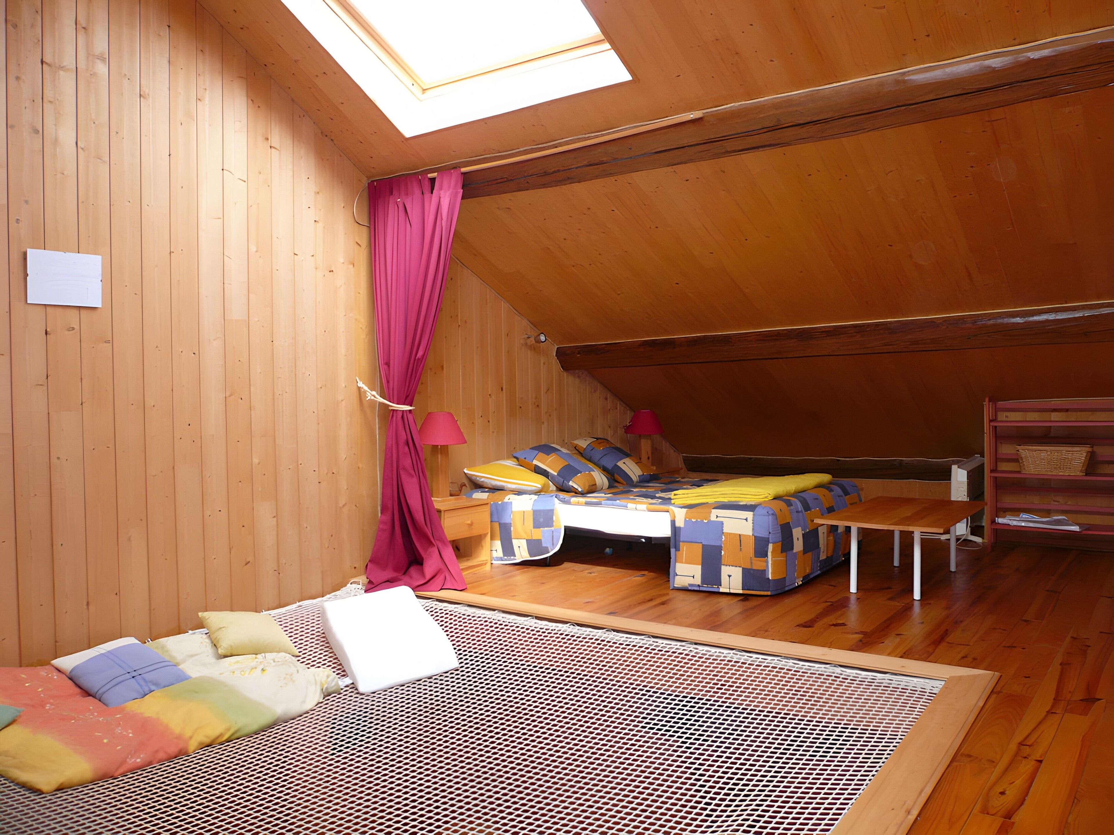 Quarto em estilo loft com teto inclinado, paredes de madeira, cama com colcha estampada, uma mesinha e uma prateleira com cesta.