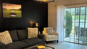 Una sala de estar moderna con un sofá oscuro, una mesa de centro de vidrio con velas, una lámpara y un cuadro de un campo de golf.