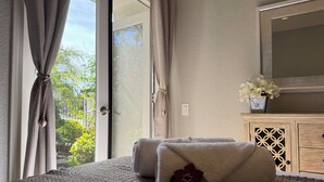 A bedroom with a large window, a bed with a textured blanket, a decorative pillow, and a small vase with flowers on a wooden cabinet. - Elegant Designer Suite: Serene Garden View & Private Entry (Escondido)