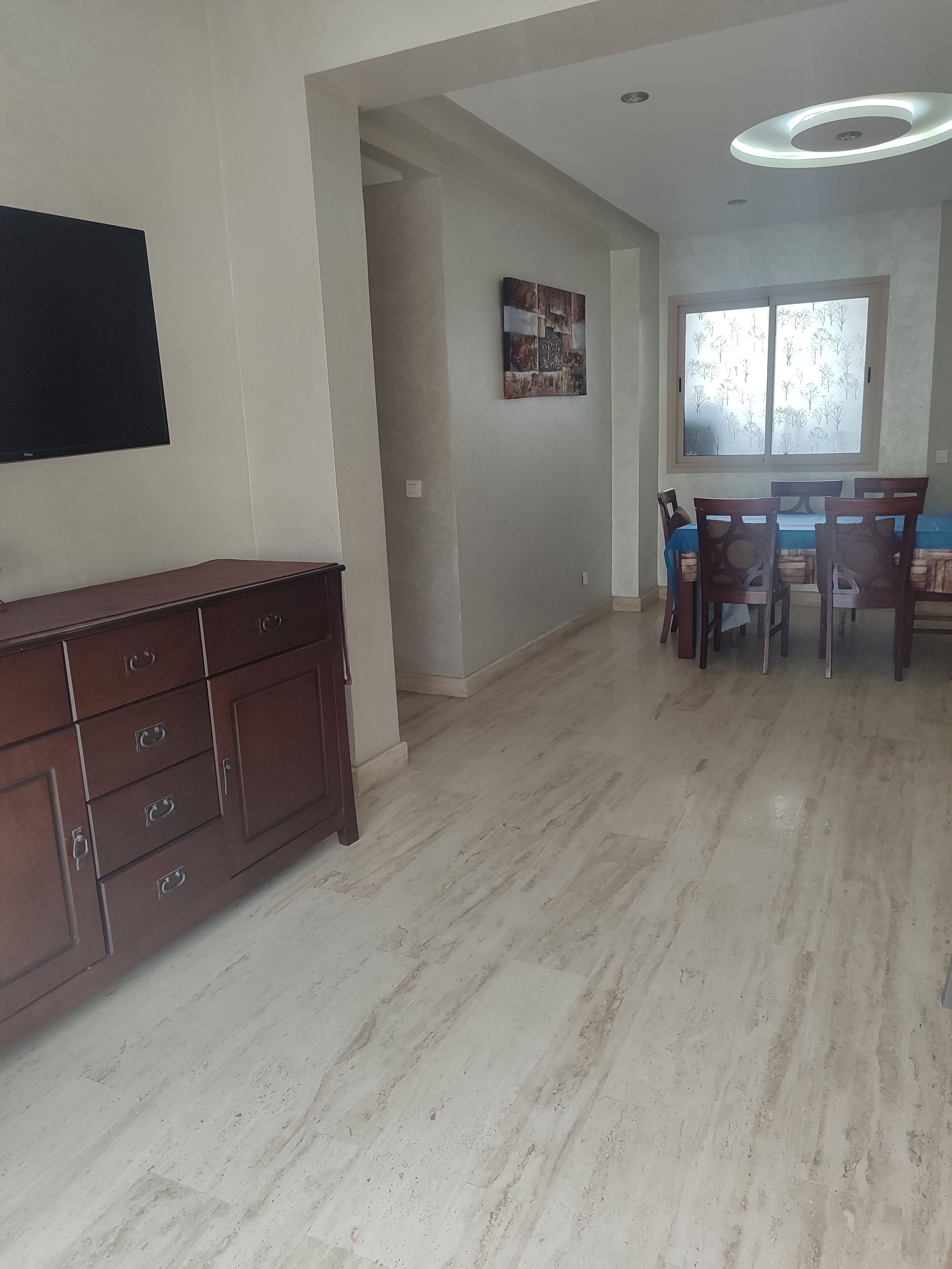 A spacious room with a wooden cabinet, a dining table with chairs, and a large window with a view of trees.