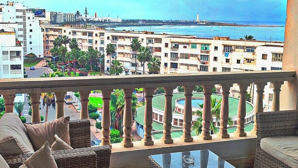 A balcony with wicker furniture, a glass coffee table, and a view of a coastal cityscape. - Melliber A  (Casablanca)