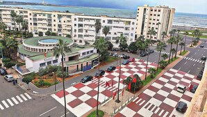 A coastal urban area with a prominent building featuring a circular rooftop pool, adjacent to a road with a checkered pedestrian crossing. - Melliber A  (Casablanca)