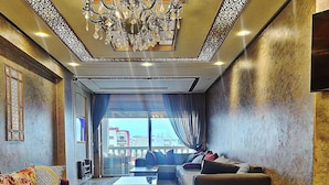 A spacious living room with a red sofa, a wooden coffee table, and a black ottoman. A chandelier hangs from the ceiling, and there is a large window with curtains. - Melliber A  (Casablanca)