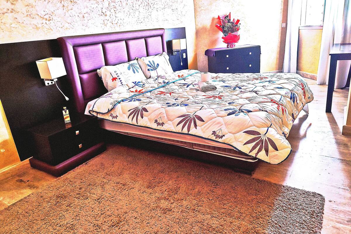 Un dormitorio con una cama grande, una mesita de noche, una lámpara y un jarrón de flores.