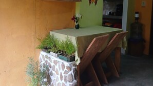 Ein Essbereich mit Tisch, Stühlen, einem Steinsockel, Topfpflanzen, einem Regal mit Büchern und Dekoration sowie einer Wand mit einem Gemälde.