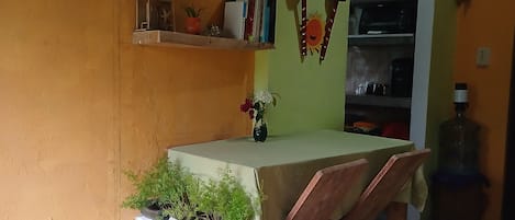 A dining area with a table, chairs, a stone base, potted plants, a shelf with books and decorative items, and a wall with a painting.