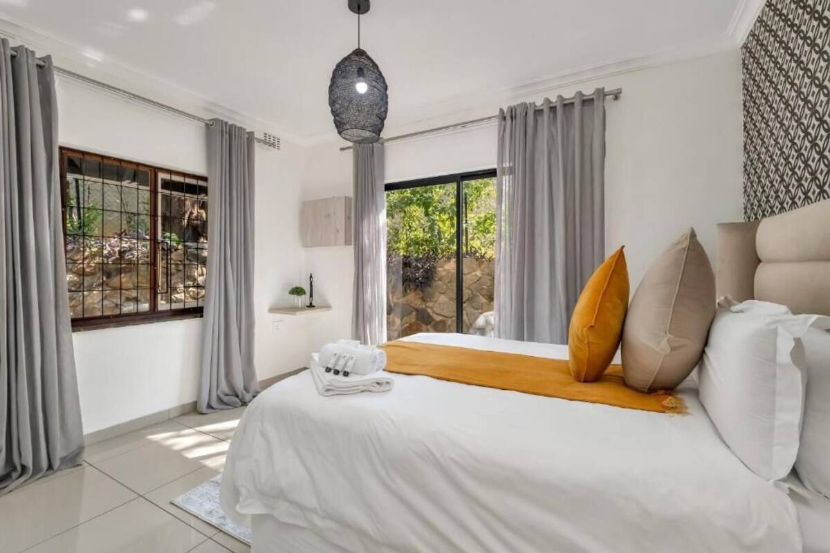 A modern hotel room with a large bed, grey curtains, a hanging light fixture, and a view of greenery outside.