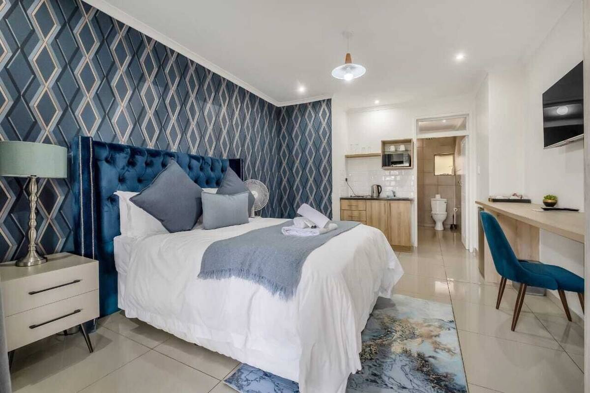 A modern bedroom with a tufted headboard, a bedside table with a lamp, a large bed with a white and gray blanket, a blue chair, and a view into the adjacent bathroom and kitchen area.