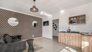 Una sala de estar moderna con un sofá gris, una mesa de centro redonda y un papel tapiz estampado. Se ve un área de cocina con armarios de madera y un salpicadero de azulejos al fondo.