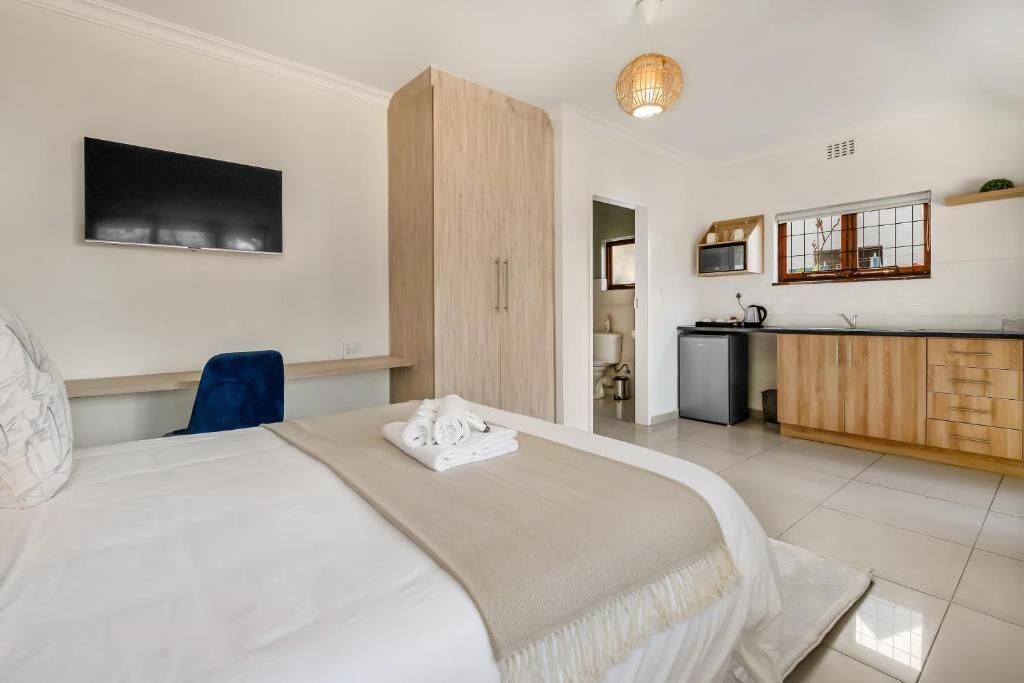 Quarto de hotel com cama, escrivaninha, guarda-roupa, cozinha compacta e televisão na parede.