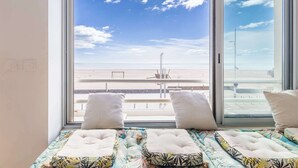 Ein Bett mit einem gemusterten Überwurf und Kissen, ein großes Fenster mit Blick auf einen Strand und eine Straße.