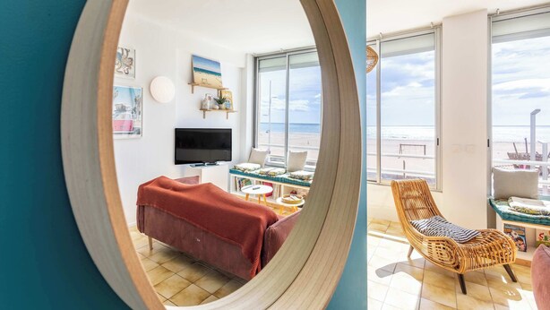 Ein Zimmer mit einem großen Spiegel, der einen Wohnbereich, einen Fernseher, eine Couch und einen Korbstuhl reflektiert, sowie Meerblick durch große Fenster.