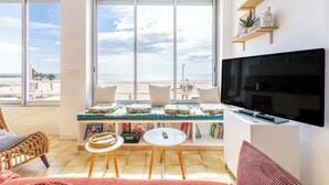 Ein Wohnzimmer mit einem großen Fernseher, einem Sofa, einem kleinen Tisch und einem Fenster mit Blick auf den Strand.