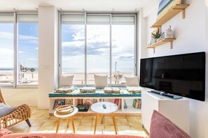 Un saló amb un televisor gran, un sofà, una taula petita i una finestra amb vista a la platja.