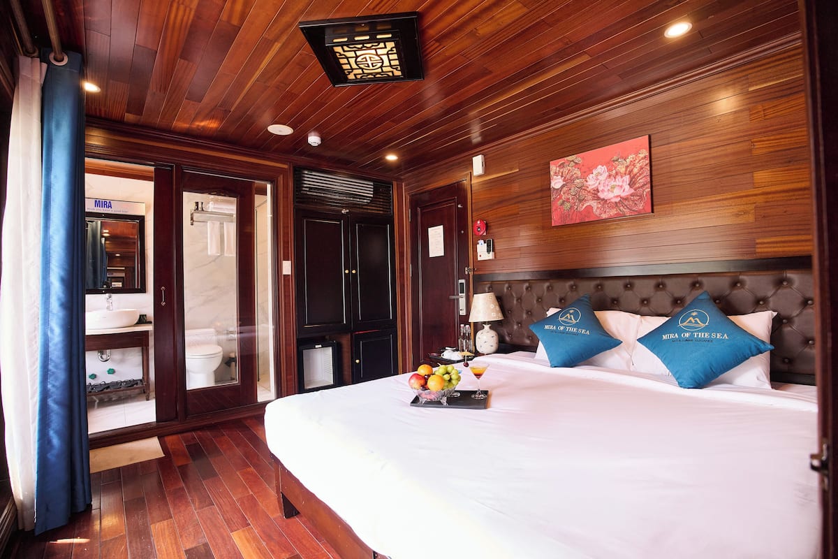 Una habitación de hotel con techo de madera, una cama con almohadas azules, un frutero en la cama y un baño con lavamanos e inodoro.