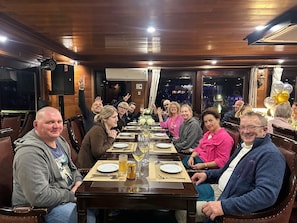 A group of people seated around a long wooden table in a restaurant with a view of the city through large windows. - Mira of the Sea Cruise Lan Ha Bay (Ha Long)