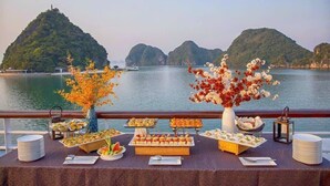 Ein Buffet mit verschiedenen Speisen und Blumenarrangements, vor einer Kulisse aus Wasser und Bergen.