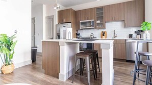 A modern kitchen with wooden cabinets, a central island with stools, stainless steel appliances, and pendant lighting. - Boulevard Stays Dashing King 1 BR Suite Pool, Parking Gym M (Richardson)