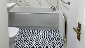 Um banheiro com uma banheira branca, uma janela pequena, um chuveiro e um vaso sanitário.