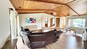 Ein geräumiger Wohnbereich mit Holzboden, einem großen Ledersofa, einem Fernseher und Blick ins Freie.