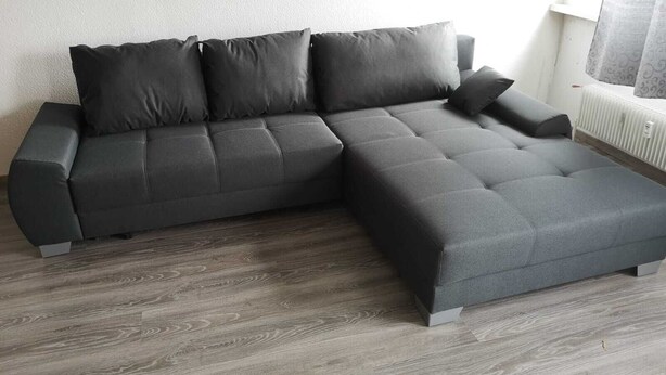 Un sofà modular en forma de L de color neutre, col·locat sobre un sòl de fusta amb un radiador blanc i una finestra amb cortina estampada al fons.