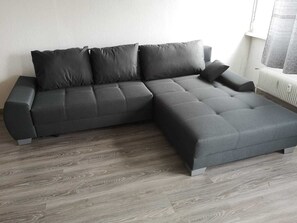 A modern L-shaped sectional sofa in a neutral color, positioned on a wooden floor with a white radiator and a window with a patterned curtain in the background.