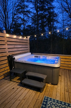 A hot tub on a wooden deck with string lights, surrounded by trees. - Blue Bird Tiny House: Hot Tub + Rooftop Deck (Logan)