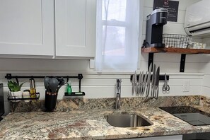 Une cuisine avec des meubles blancs, un plan de travail en granit, une machine à café et une fenêtre ornée de rideaux légers.