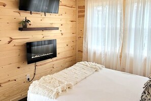 A wooden-paneled room with a bed, a TV mounted on the wall, and a window with sheer curtains. - Blue Bird Tiny House: Hot Tub + Rooftop Deck (Logan)