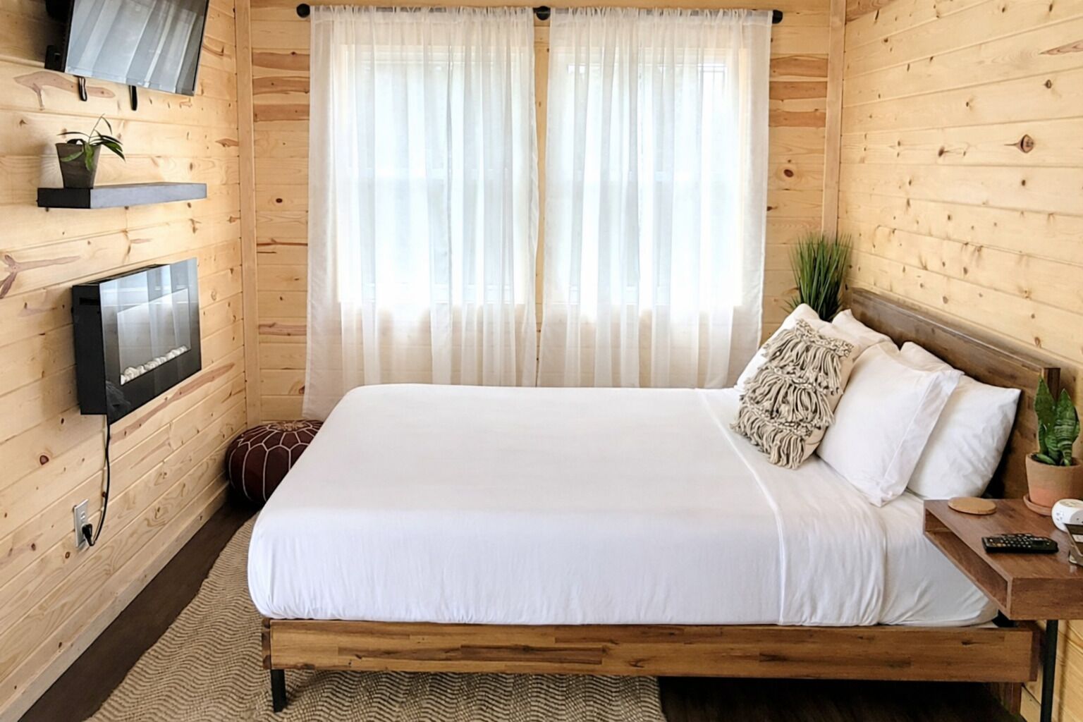 A wooden-paneled room with a bed, a mounted TV, and a window with sheer curtains.