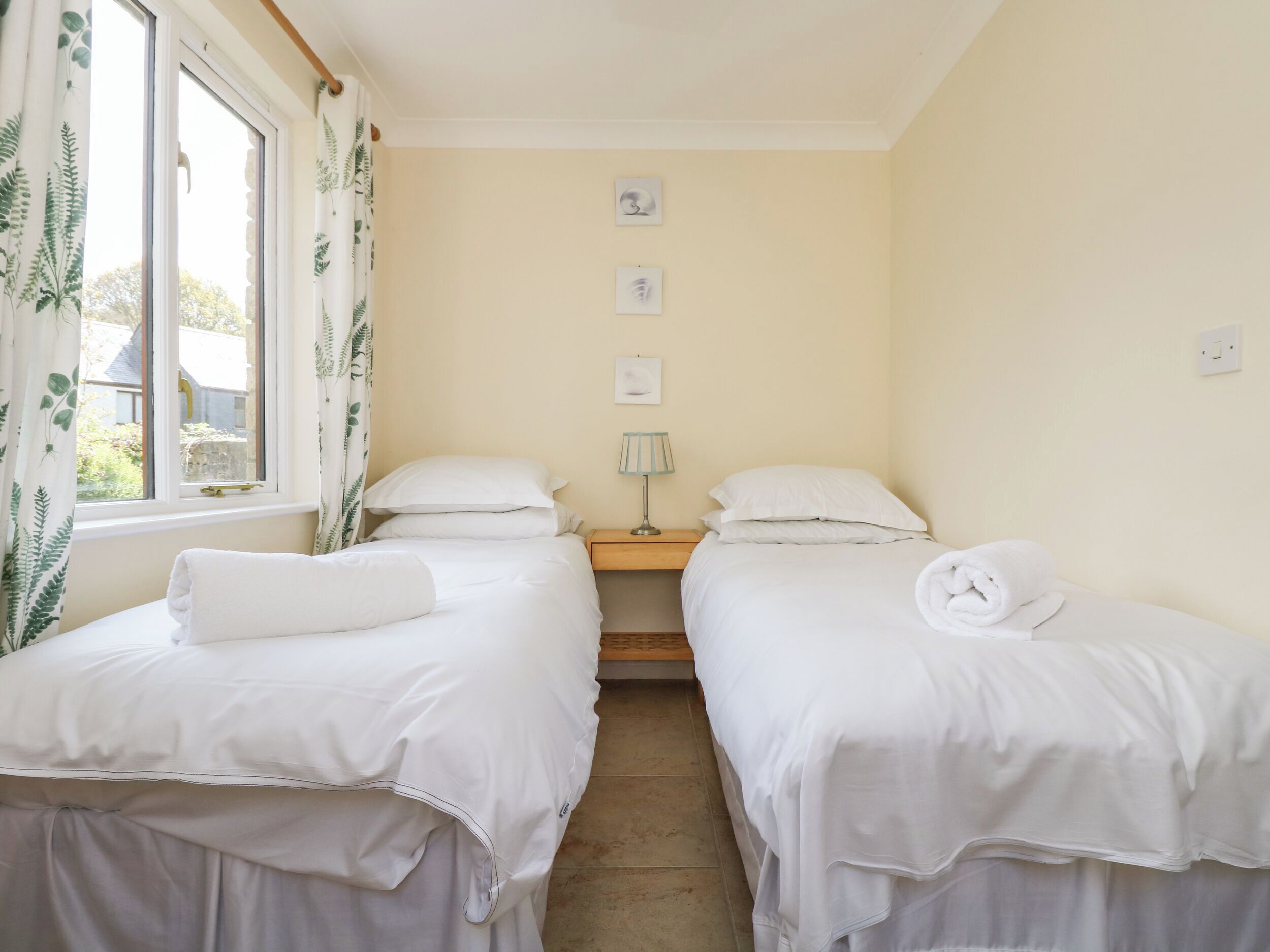 Quarto com duas camas de solteiro, mesa de cabeceira com luminária e janela com cortinas.