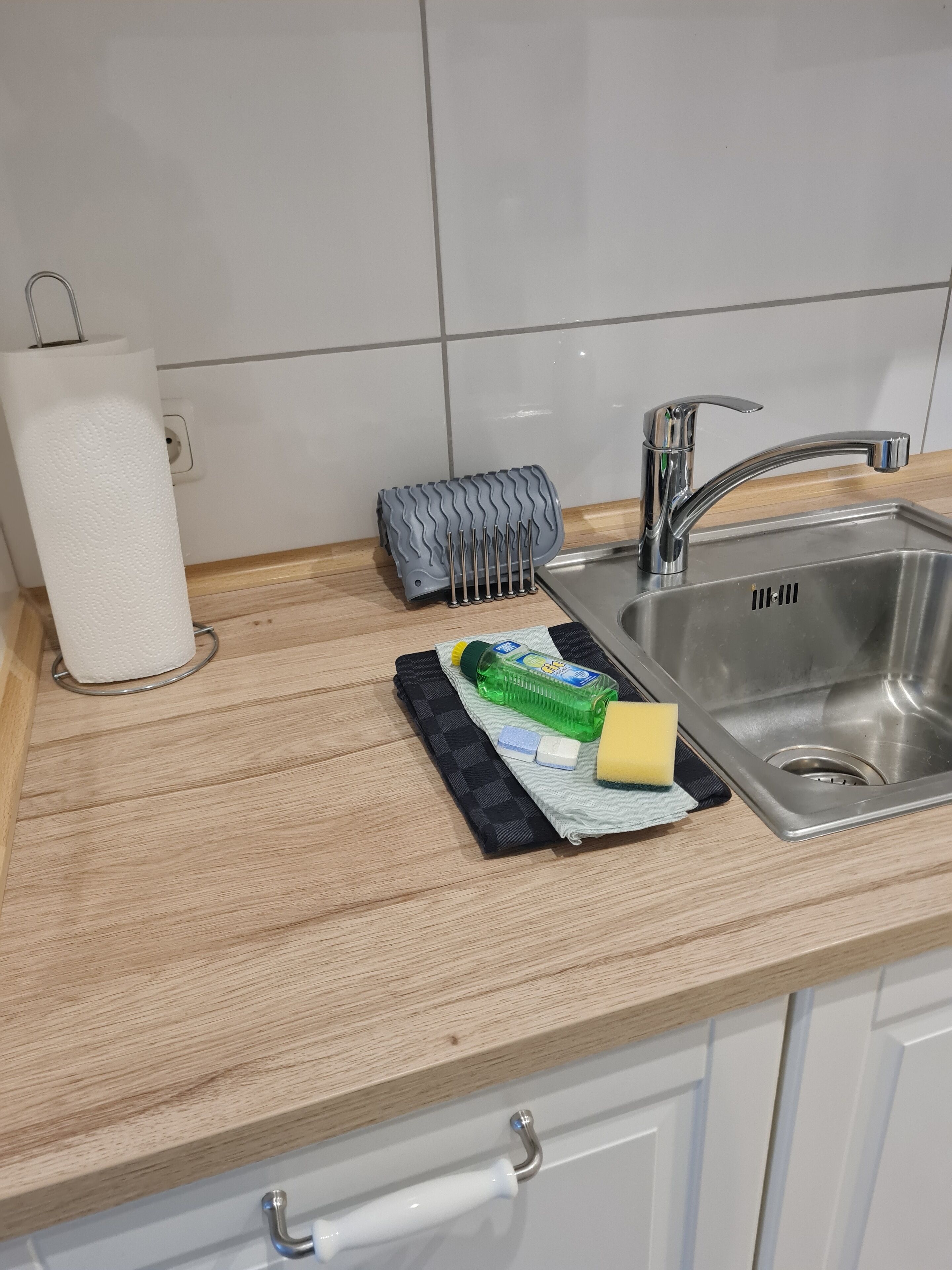 A kitchen counter with a paper towel holder, a dish rack, a bottle of dish soap, a sponge, and a dish brush.