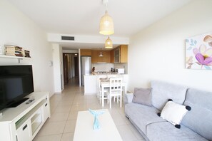 A modern living room with a flat-screen TV, a white coffee table with a blue object, a grey sofa with a white pillow, and a kitchen in the background. - Apartment Panoramic B2 by Parc Mont-roig (Miami Playa)
