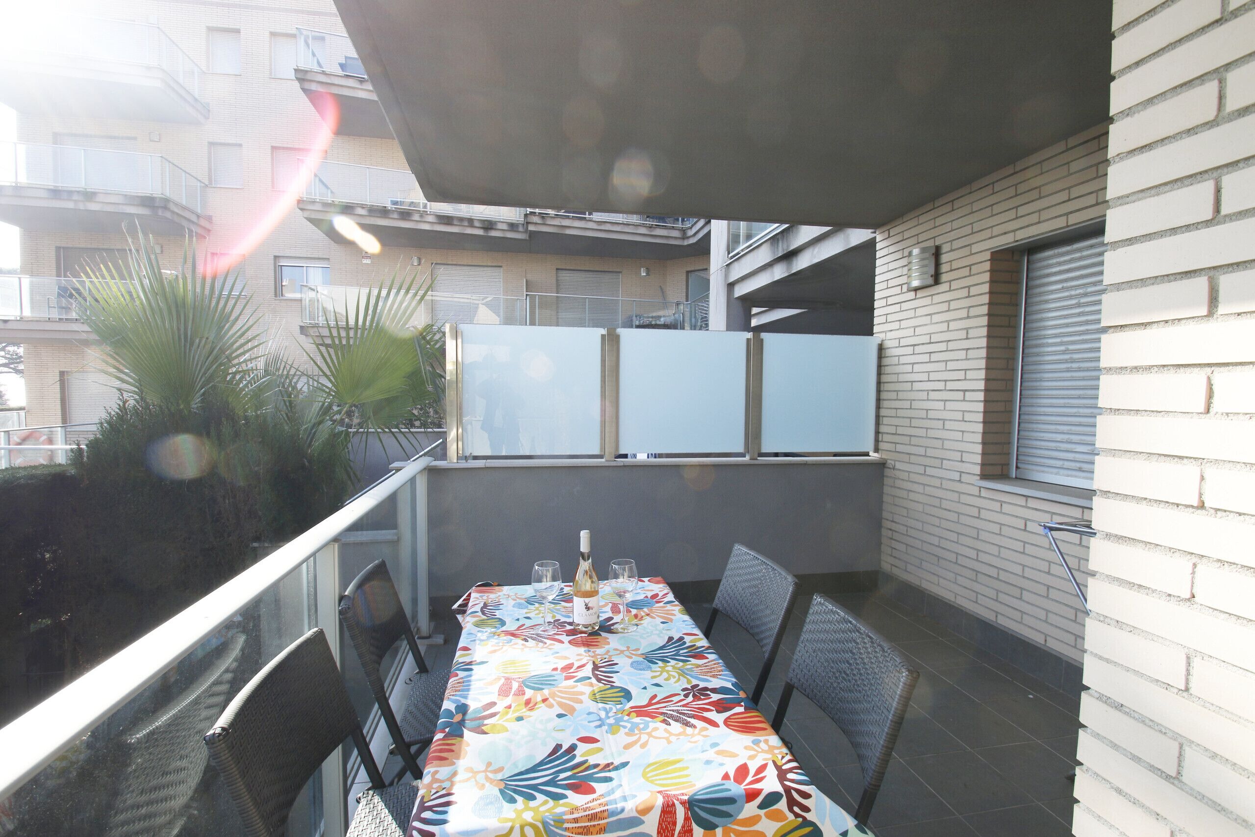 Un balcón con una mesa dispuesta para dos personas, con un mantel colorido y una botella de vino.