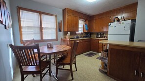 Une cuisine avec des meubles de rangement en bois, une table à manger avec des chaises, un réfrigérateur et une fenêtre avec des stores.
