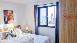 A hotel room with a bed, a nightstand, a lamp, a window with curtains, and a painting on the wall. - Sandine House (Estreito da Calheta)