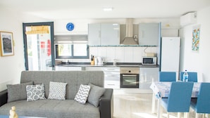 A modern living room with a grey sofa, a small table, and a dining area with blue chairs. - Sandine House (Estreito da Calheta)