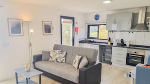 A modern living room with a grey sofa, a small table, and a kitchen area with cabinets, a stove, and a sink. - Sandine House (Estreito da Calheta)