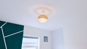 Une pièce avec un luminaire au plafond, une fenêtre avec des stores et un tableau encadré accroché au mur.