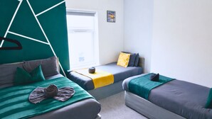 Une chambre avec deux lits : l’un est recouvert d’une couverture et de coussins à rayures vertes et grises, et l’autre d’une couverture et de coussins bleus et gris. Il y a une fenêtre donnant sur l’extérieur, une petite table avec un coussin jaune et un tableau accroché au mur.