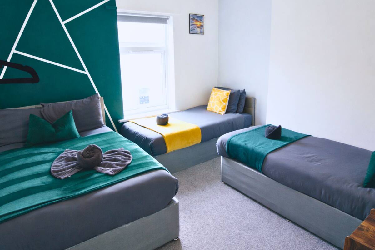 A room with two beds, one with a green and gray striped blanket and pillows, and the other with a blue and gray blanket and pillows. There is a window with a view of the outside, a small table with a yellow cushion, and a picture on the wall.
