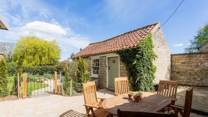 Une petite maison en pierre avec un toit en tuiles, une table en bois avec des chaises et un petit jardin.