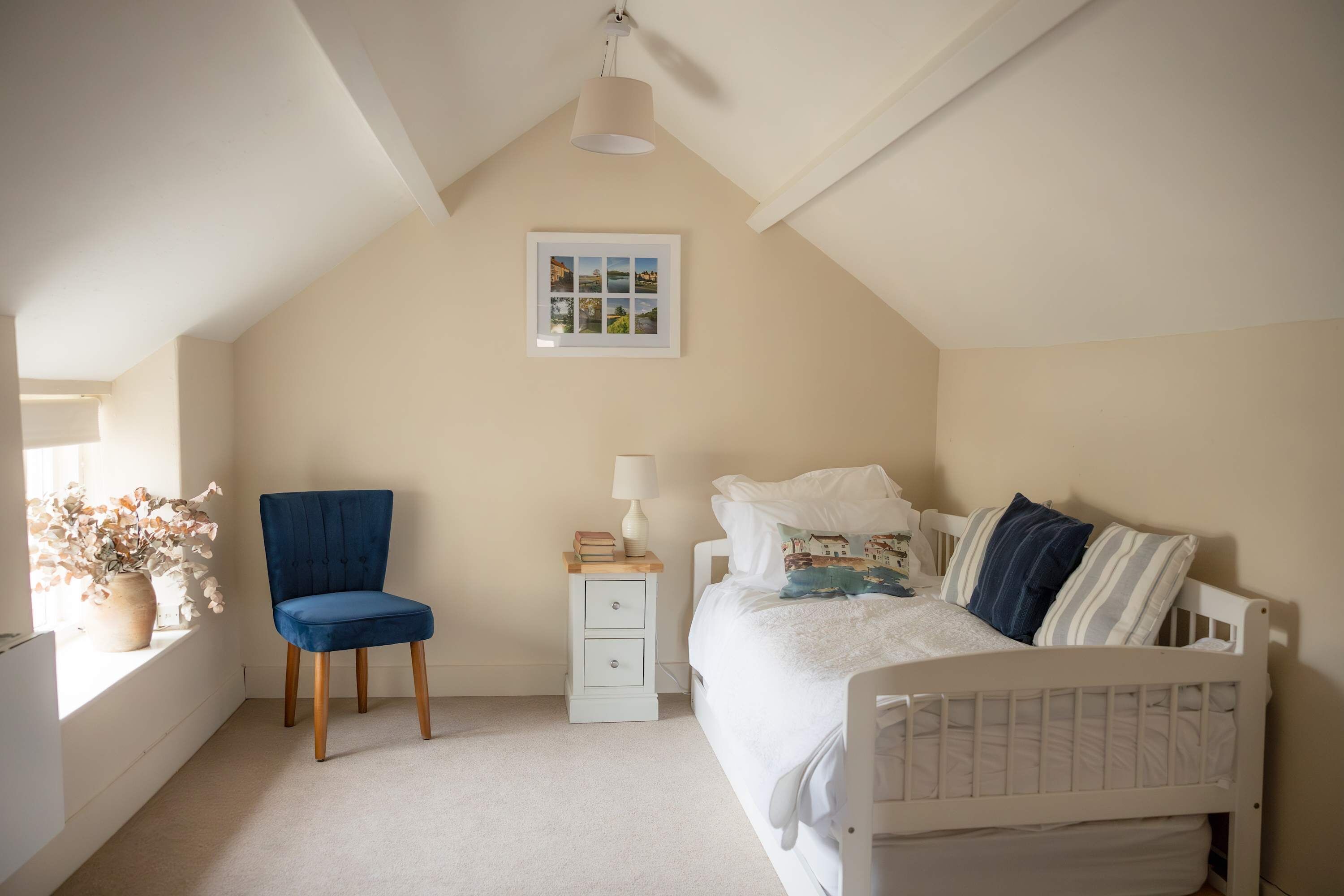 A bedroom with a sloped ceiling, a bed with white bedding, a blue chair, a bedside table with a lamp, and a framed photo on the wall.