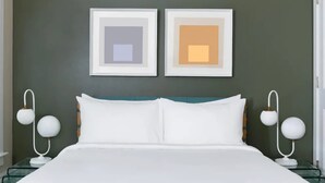 Un lit bien fait, avec du linge de lit blanc, deux tables de chevet avec des lampes blanches, et deux œuvres d’art encadrées accrochées au mur.