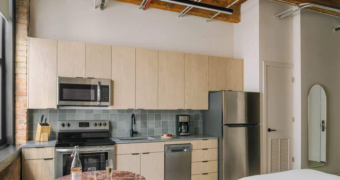 A modern kitchen with light wood cabinets, a stainless steel microwave, oven, and refrigerator. There is a sink with a faucet, a coffee maker, and a wine bottle on the counter.