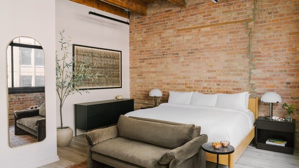 A modern bedroom with a brick wall, a large bed, a sofa, a nightstand, and a mirror. - LIVN West Loop Chicago King Studio (Chicago)