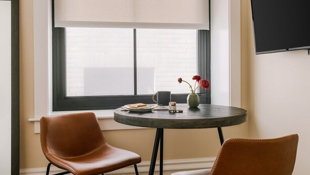 A room with a round table, two chairs, a vase with flowers, a plate with food, and a window with blinds.