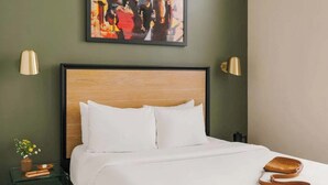 A hotel room with a wooden headboard, white bedding, a small bedside table with a vase of flowers, and a brown leather bag on the bed.